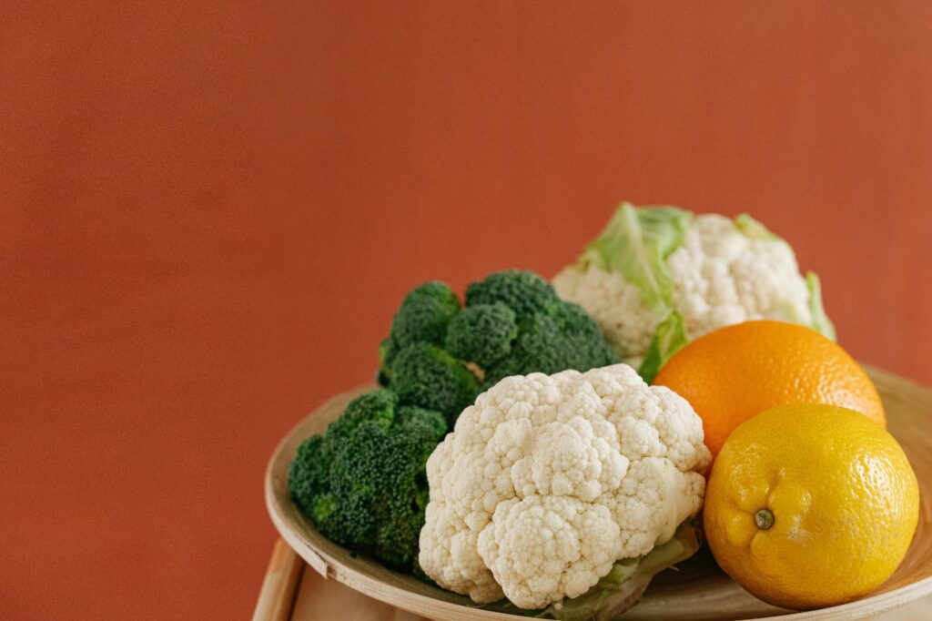 Close-up of fresh broccoli, cauliflower, lemon, and orange on a wooden plate with an earthy background.
